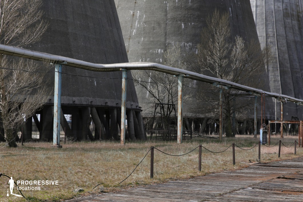 This vast industrial landscape features massive concrete cooling towers rising in the background, their weathered surfaces showing signs of age and exposure. A network of rusted, elevated pipelines runs across the foreground, supported by corroded metal columns that add to the scene’s sense of decay. The cracked concrete pathways and overgrown grass contrast with the towering industrial structures, reinforcing the abandoned and desolate atmosphere. This location is ideal for dystopian, sci-fi, or post-apocalyptic films, providing an imposing and visually striking setting for themes of industrial decline, secrecy, or environmental collapse.