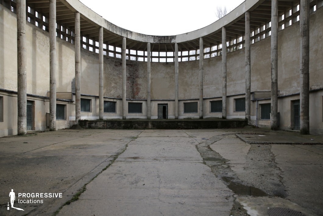 This imposing, semi-circular structure features a rhythmic row of tall, weathered concrete columns supporting an open-air upper level with rectangular cutouts, allowing diffused light to filter through. The lower section consists of small, barred windows and aged plaster walls, marked by peeling paint and patches of exposed brick, adding to the sense of decay. The cracked concrete ground, with visible water damage and overgrown cracks, reinforces the abandoned and austere atmosphere. This location is ideal for dystopian, historical, or thriller productions, serving as a forgotten institutional courtyard, a prison yard, or a brutalist architectural relic.