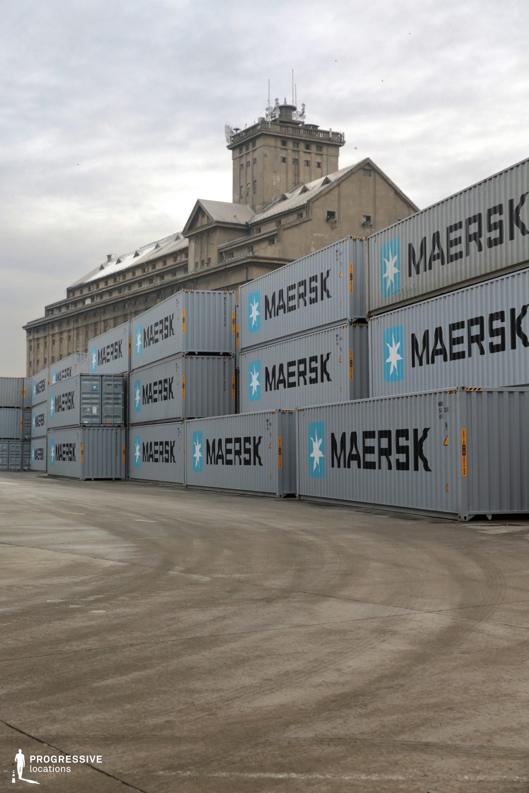 This industrial setting features a neatly stacked arrangement of shipping containers in the foreground, creating a structured and repetitive pattern. The towering historic concrete silo in the background contrasts with the modern logistics operation, adding depth and a sense of industrial heritage to the scene. The expansive concrete ground, marked with tire tracks, reinforces the location’s functionality, while the muted, overcast sky enhances the gritty, utilitarian atmosphere. This site is ideal for filming crime thrillers, action sequences, or dystopian narratives, providing a dynamic and imposing industrial backdrop for high-stakes storytelling.