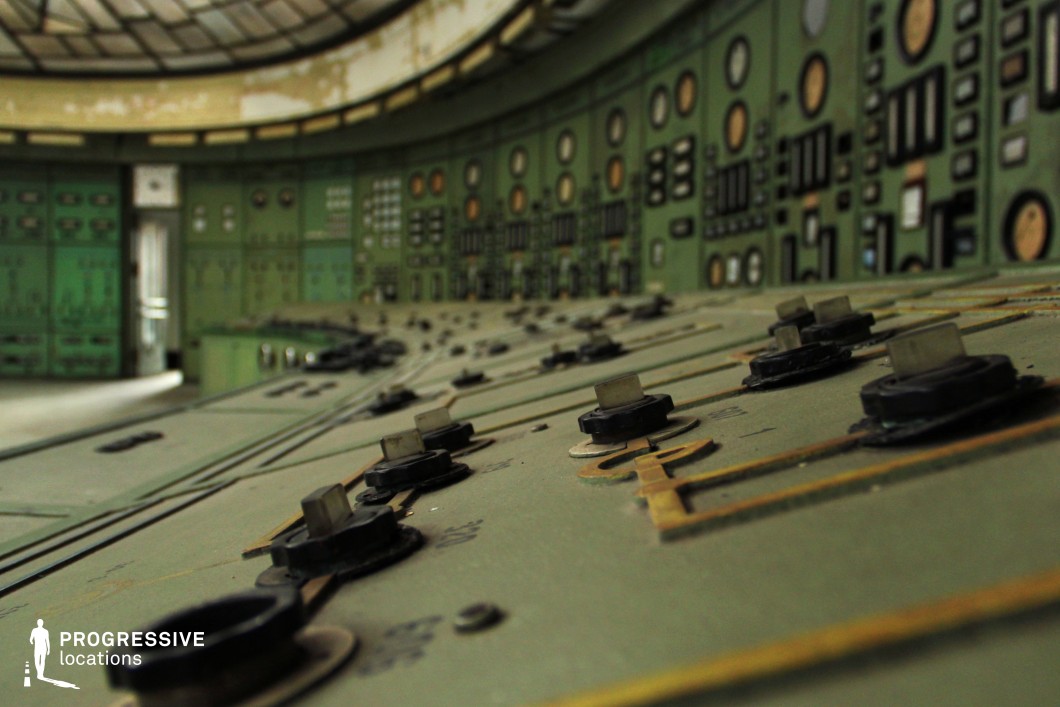 This detailed close-up captures an aged industrial control panel with a textured green metal surface, covered in an array of black rotary switches, buttons, and engraved circuit lines. The worn edges and slightly corroded elements indicate long-term disuse, reinforcing a vintage, utilitarian aesthetic. The shallow depth of field emphasizes the tactile nature of the controls while leading the eye toward the background of dials and meters on the curved wall. This visually rich setting is ideal for sci-fi, dystopian, or historical thriller productions, serving as an abandoned power facility, a secret control center, or a retro-futuristic command station.