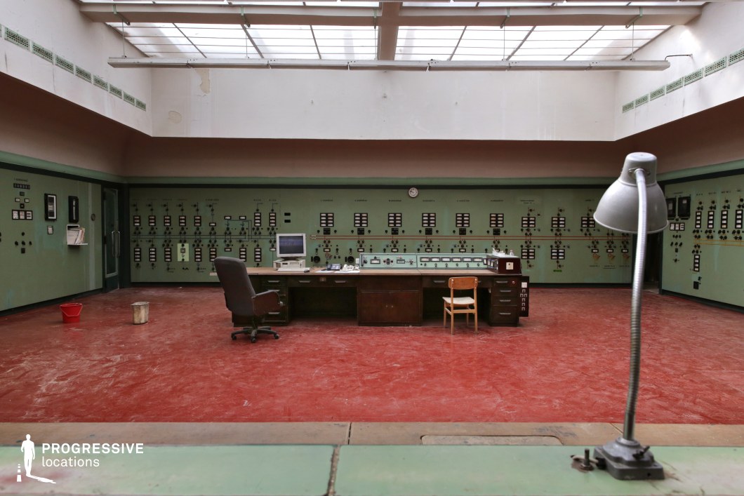 This vintage industrial control room features a striking red floor that contrasts sharply with the muted green metal control panels lining the walls, enhancing the space’s utilitarian yet visually distinct aesthetic. The symmetrical design is anchored by a central wooden desk, complete with an old computer and analog equipment, reinforcing a mid-to-late 20th-century operational setting. Overhead, a large skylight casts diffused natural light, highlighting the faded textures and worn industrial elements. The interplay of bold colors and structured symmetry makes this an ideal location for thriller, sci-fi, or historical productions, serving as an abandoned power station control center, a classified research facility, or a Cold War-era command post.