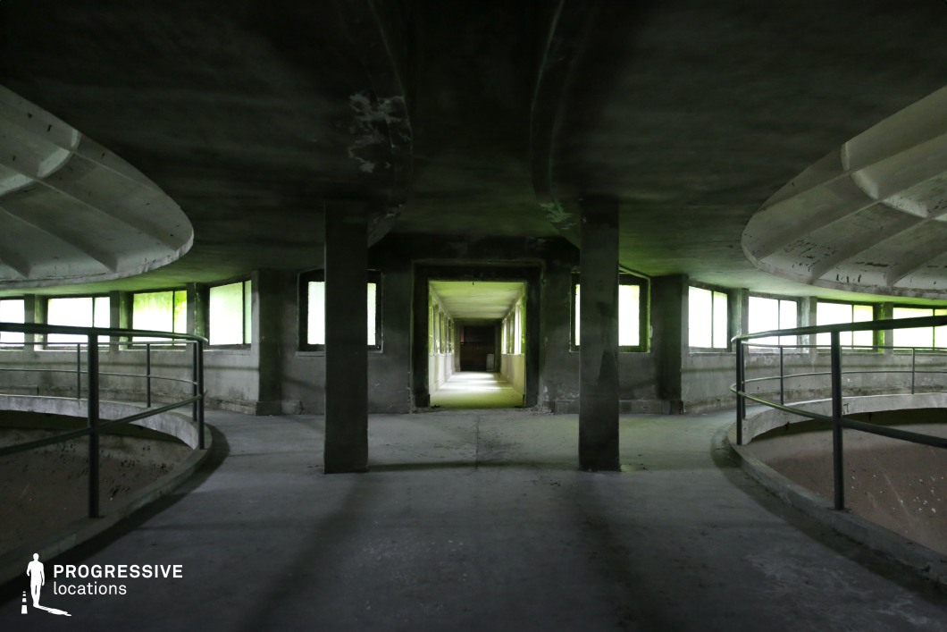 This circular industrial space features a raw concrete structure with an open, layered design. A curved walkway with metal railings encircles the space, flanked by a series of evenly spaced rectangular windows that cast an eerie greenish light across the surfaces. Thick concrete beams and columns divide the area, leading towards a long, dimly lit corridor that extends into the distance, creating a sense of depth and mystery. The stark geometry and brutalist design make this location ideal for filming dystopian, sci-fi, or psychological thriller scenes, providing a futuristic yet decayed setting with a strong atmospheric presence.