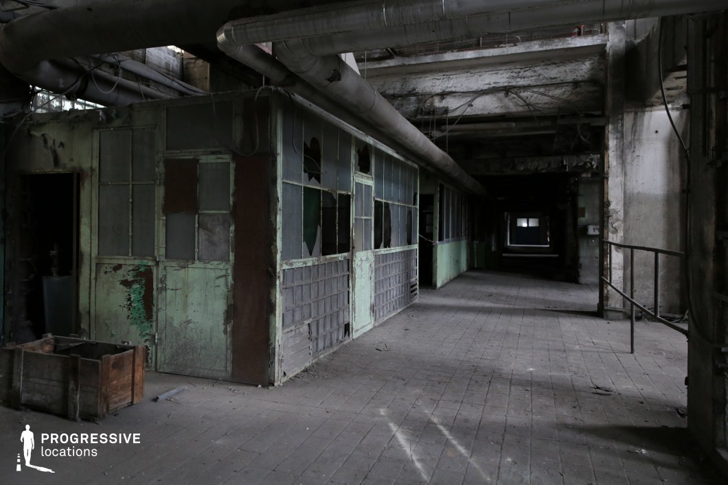 This abandoned industrial corridor features cracked and peeling green walls, shattered glass panels, and exposed metal frameworks, creating an atmosphere of neglect and decay. Overhead, large industrial pipes snake through the ceiling, while corroded beams and exposed wiring add to the raw, unkempt aesthetic. The dimly lit passage extends into darkness, with scattered debris and worn tiled flooring emphasizing its derelict state. This location is ideal for filming psychological thrillers, dystopian, or horror scenes, providing a visually rich setting for tension, suspense, and atmospheric storytelling.