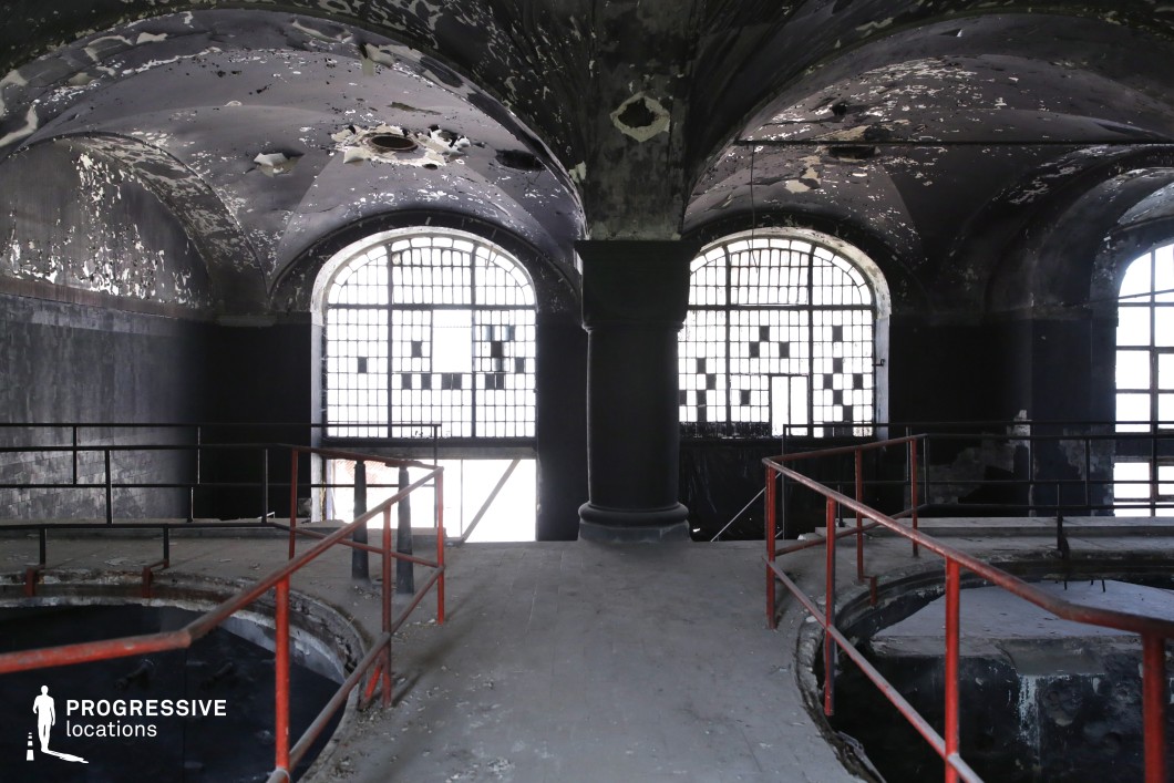 This abandoned industrial space features towering arched windows with fragmented glass, casting a diffused glow into the dimly lit interior. The vaulted ceiling, marked by peeling paint and exposed patches of decay, adds a haunting, gothic atmosphere. A raised walkway with metal railings leads toward the center, flanked by deep circular openings that suggest past mechanical or industrial use. The stark contrast between light and shadow, along with the grand yet deteriorated architecture, makes this location ideal for horror, dystopian, or neo-noir productions, serving as an abandoned power station, a secret underground chamber, or a forgotten historical site.