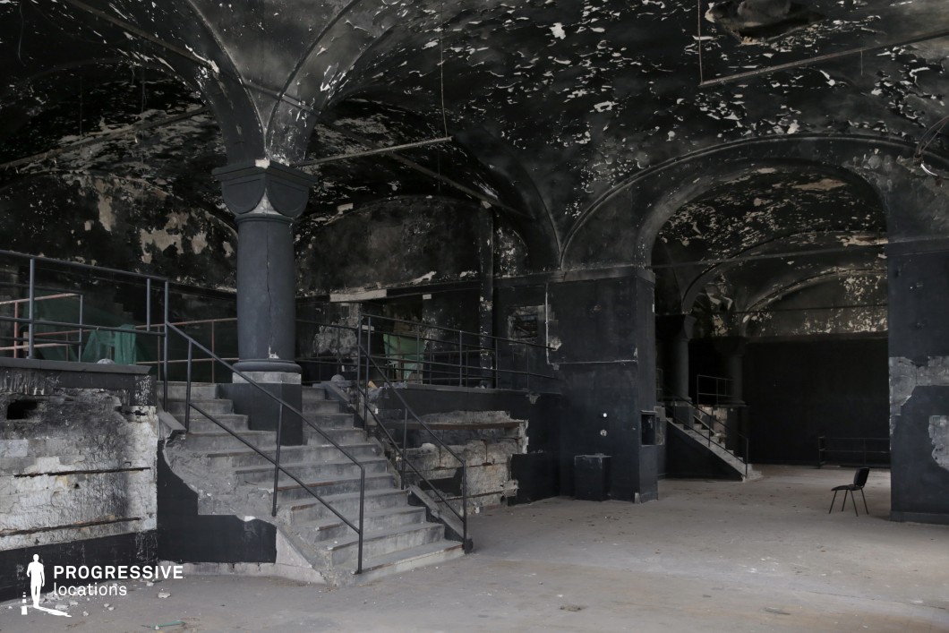 This abandoned interior features high, vaulted ceilings supported by thick, dark columns, creating a cavernous and imposing atmosphere. The walls and ceiling are heavily weathered, with peeling blackened paint and exposed patches of aged plaster, giving the space a distressed and timeworn look. A grand staircase with metal railings leads to an elevated platform, while additional staircases and open walkways suggest a once-functional multi-level structure. The vast open floor, scattered debris, and stark contrast between shadowy recesses and ambient light contribute to an eerie, post-apocalyptic aesthetic. This location is ideal for filming dark, atmospheric scenes, such as horror, dystopian thrillers, or crime dramas, offering a raw and cinematic backdrop for suspenseful storytelling.