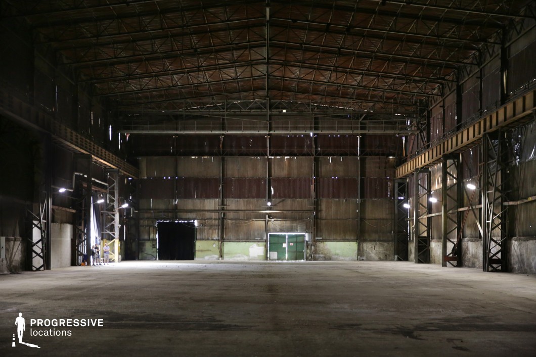 This expansive, dimly lit warehouse features towering steel framework, a rusted corrugated metal facade, and an immense open floor space. The high ceiling, supported by a skeletal metal truss structure, adds a sense of scale, while industrial lighting casts long shadows across the worn concrete surface. The weathered walls, punctuated by patches of peeling paint and rust, contribute to the abandoned and gritty atmosphere. Large doors at the far end suggest former heavy-duty industrial use. This location is ideal for filming action, dystopian, or crime sequences, offering a dramatic setting for high-stakes confrontations or atmospheric, suspenseful storytelling.