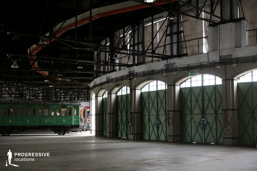 This industrial train depot features a curved row of arched doors with large green metal gates, framed by textured concrete and exposed steel beams supporting the high ceiling. The structure exhibits a combination of functional industrial architecture and historic design elements. Inside, vintage green and wooden railway carriages rest on the tracks, adding a nostalgic atmosphere. Overhead, suspended lights and metal framework create depth, while natural light filtering through the arched windows enhances the aged aesthetic. This location is ideal for filming period dramas, historical adventure films, or noir-style mysteries, providing a cinematic backdrop for scenes involving rail transport, travel, or industrial heritage.