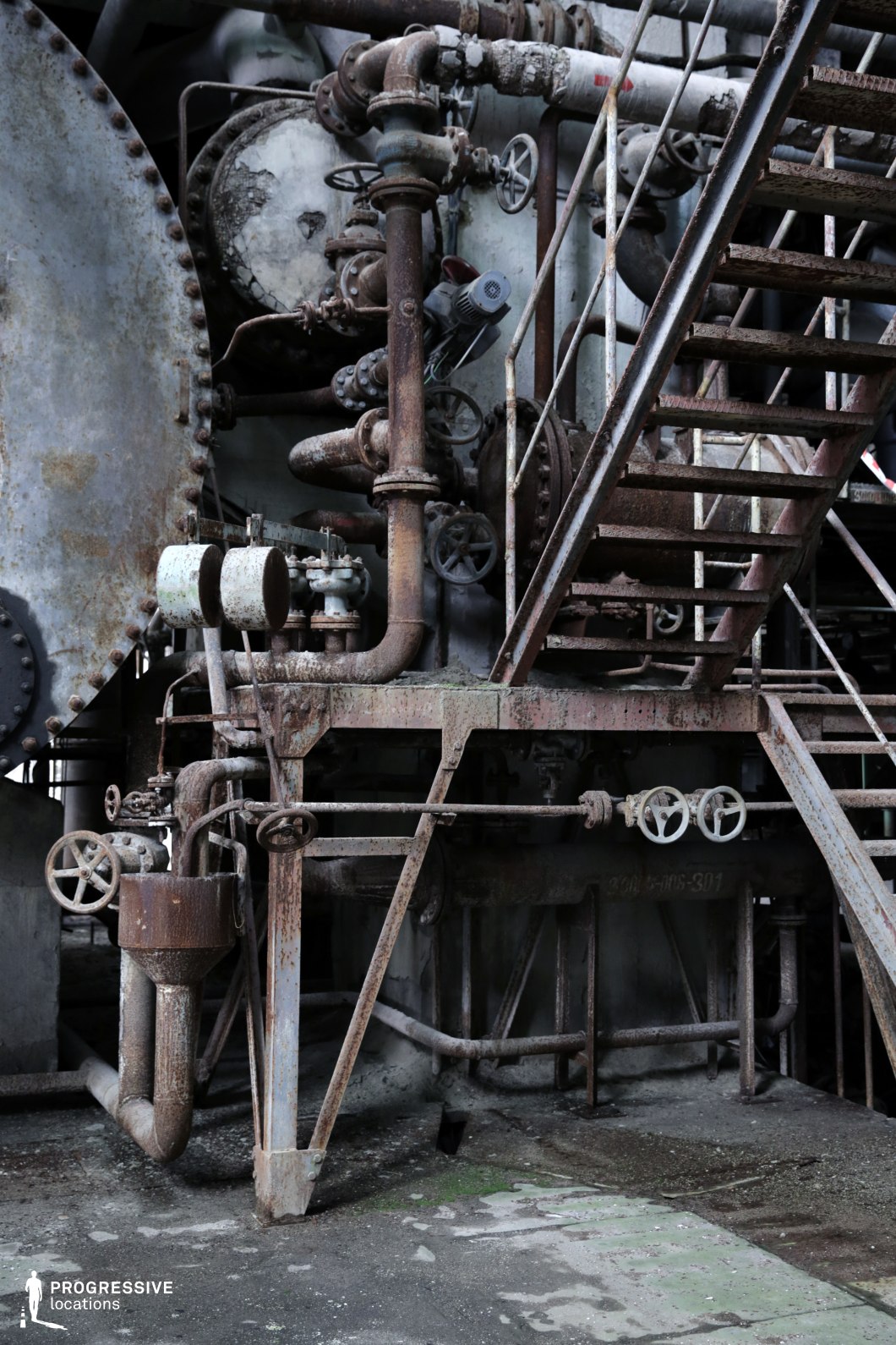 This close-up view of an aged industrial facility features a dense network of corroded pipes, valves, and metal staircases, emphasizing a raw and mechanical aesthetic. The rusted surfaces, peeling paint, and worn textures highlight the passage of time and abandonment. Large riveted metal tanks and interconnected tubing add to the industrial complexity, while the stairway provides a sense of scale and verticality. This location is ideal for filming dystopian, steampunk, or cyberpunk-themed scenes, as well as industrial crime dramas or action sequences that require a gritty, atmospheric backdrop.