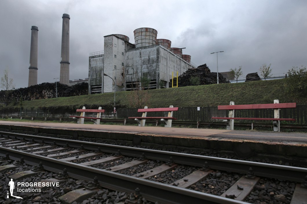 This location features an aging railway platform with red and white benches overlooking a set of steel train tracks in the foreground. A weathered industrial facility with large cylindrical cooling towers and towering chimneys dominates the background, its corroded metal panels and exposed brick suggesting decades of operation and decay. The raised grassy embankment with fencing and scattered vegetation adds a subtle contrast to the industrial severity. The combination of railway infrastructure and abandoned factory aesthetics makes this an ideal setting for thriller, dystopian, or noir films, serving as a remote train stop, an industrial-era transit hub, or a post-apocalyptic wasteland.