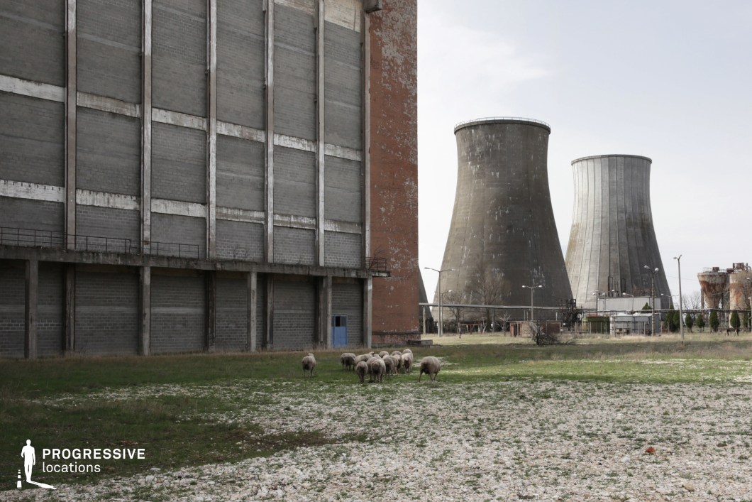 This expansive industrial setting features a large, weathered concrete structure with towering cooling towers in the background, evoking a sense of scale and industrial decline. The foreground consists of a rough, patchy field with scattered stones, contrasting with the rigid geometric facades of the factory buildings. Rusted metal details and peeling surfaces enhance the sense of abandonment. The stark, functional architecture and desolate landscape make this an ideal location for dystopian, post-apocalyptic, or thriller productions, serving as an abandoned power facility, a decayed industrial zone, or a setting for environmental and science-fiction narratives.