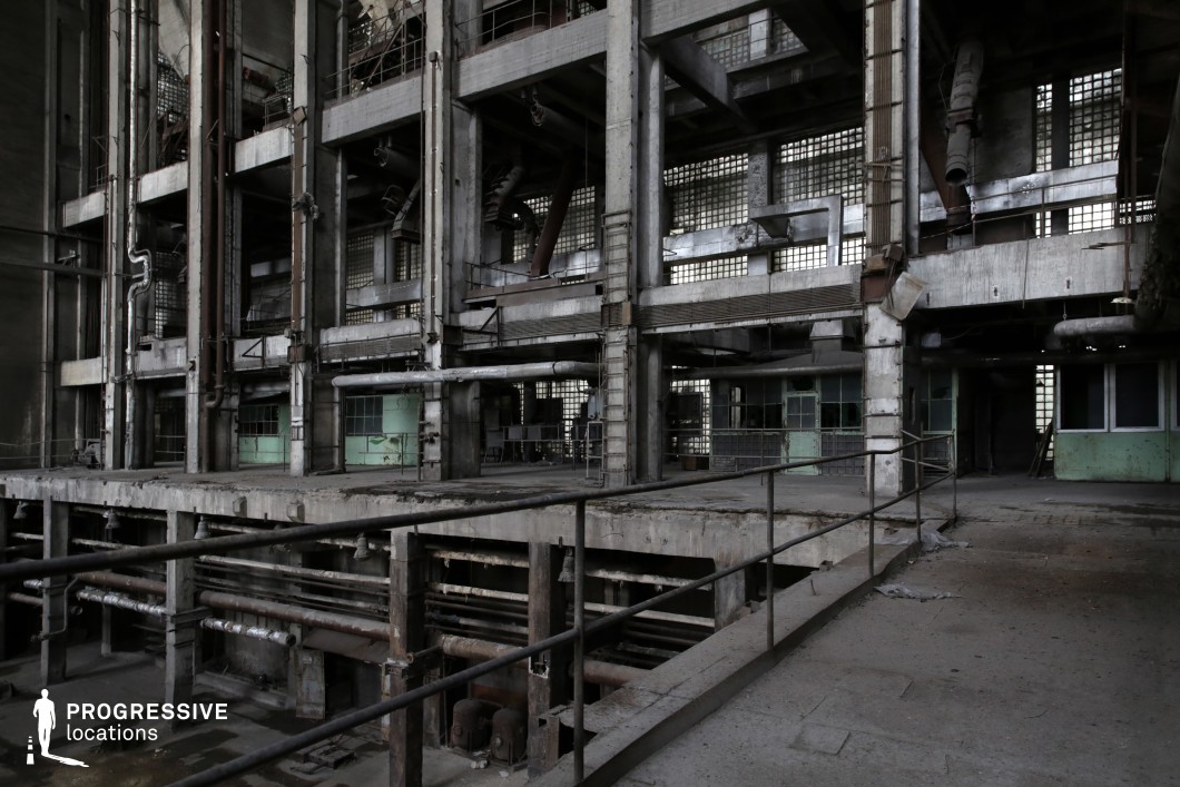 This massive, decayed industrial complex features a multi-level concrete framework lined with rusted metal pipes, exposed ducts, and broken railings. The grid-like pattern of glass block windows casts a muted, diffused light into the space, contrasting with the heavy industrial elements. The structure includes walkways, ladders, and platforms, reinforcing its former role as a production or processing facility. Crumbling surfaces and aged machinery remnants add to the sense of abandonment. This location is ideal for filming dystopian, sci-fi, or action sequences, offering a layered, mechanical backdrop perfect for chase scenes, secret experiments, or post-apocalyptic settings.