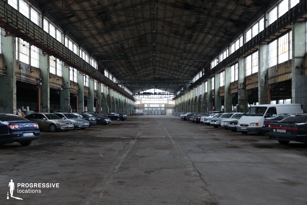 This expansive industrial hall features a towering metal roof structure supported by large, weathered concrete columns. Rows of tall, multi-paned windows line both sides, allowing diffused natural light to filter in, casting long shadows across the cracked and worn concrete floor. The rusted overhead railings and peeling paint on the walls emphasize the hall’s aged, utilitarian aesthetic. The immense open space and dramatic perspective make this location ideal for filming action sequences, dystopian thrillers, or crime dramas, offering a raw, industrial setting with a cinematic sense of depth and scale.