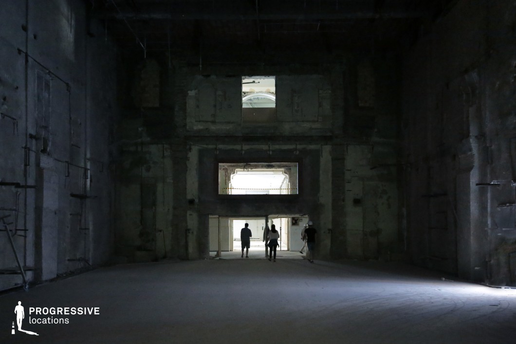 This vast, abandoned hall features towering concrete walls, exposed structural elements, and a cavernous ceiling, creating a stark and ominous atmosphere. The rough, unfinished surfaces are marked by remnants of past construction, with embedded metal rods and uneven textures adding to the sense of decay. A series of open windows and doorways in the background allow glimpses of brighter, more structured spaces, creating a strong contrast between light and shadow. This location is ideal for filming dystopian, horror, or psychological thriller scenes, offering an imposing and mysterious setting for tension-filled storytelling.