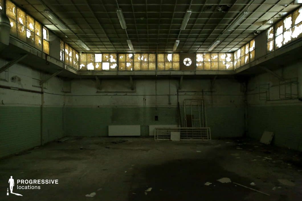 This desolate industrial hall features a high ceiling with a grid of deteriorating panels and rows of shattered upper windows that allow minimal, diffused light to seep in. The walls, partially covered in faded green tiles, contrast with the exposed pipes and peeling paint, reinforcing the sense of neglect. The cracked concrete floor is scattered with debris, while metal grates and discarded frames lean against the walls, suggesting a space long forgotten. The eerie, dimly lit atmosphere makes this an ideal filming location for horror, dystopian, or psychological thriller scenes, offering a backdrop filled with tension and decay.