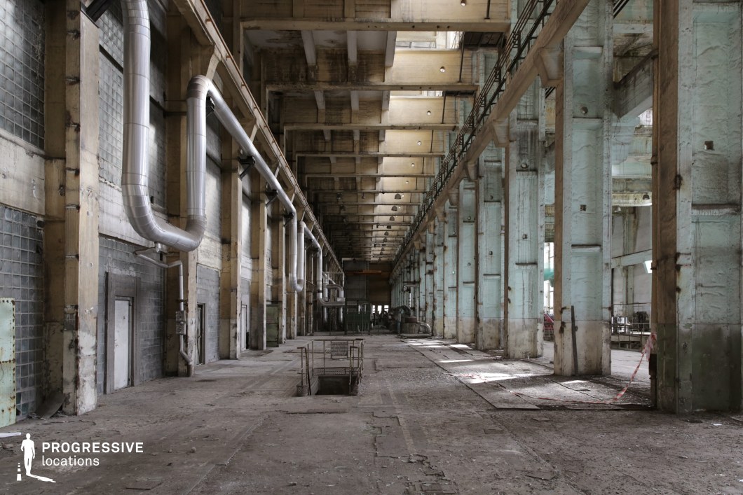 A monumental abandoned factory hall featuring towering concrete columns and a high, segmented ceiling with a repetitive structural design. The walls have faded greenish-blue paint peeling away, revealing aged concrete. Large industrial ventilation pipes run along the walls, adding to the mechanical character of the space. The floor is worn, cracked, and covered in dust, with open pits and remnants of past machinery hinting at previous heavy industrial use. The long, narrow perspective leads to distant equipment in the background, offering excellent depth for cinematography. Ideal for industrial, dystopian, or sci-fi productions requiring a grand, decayed setting.