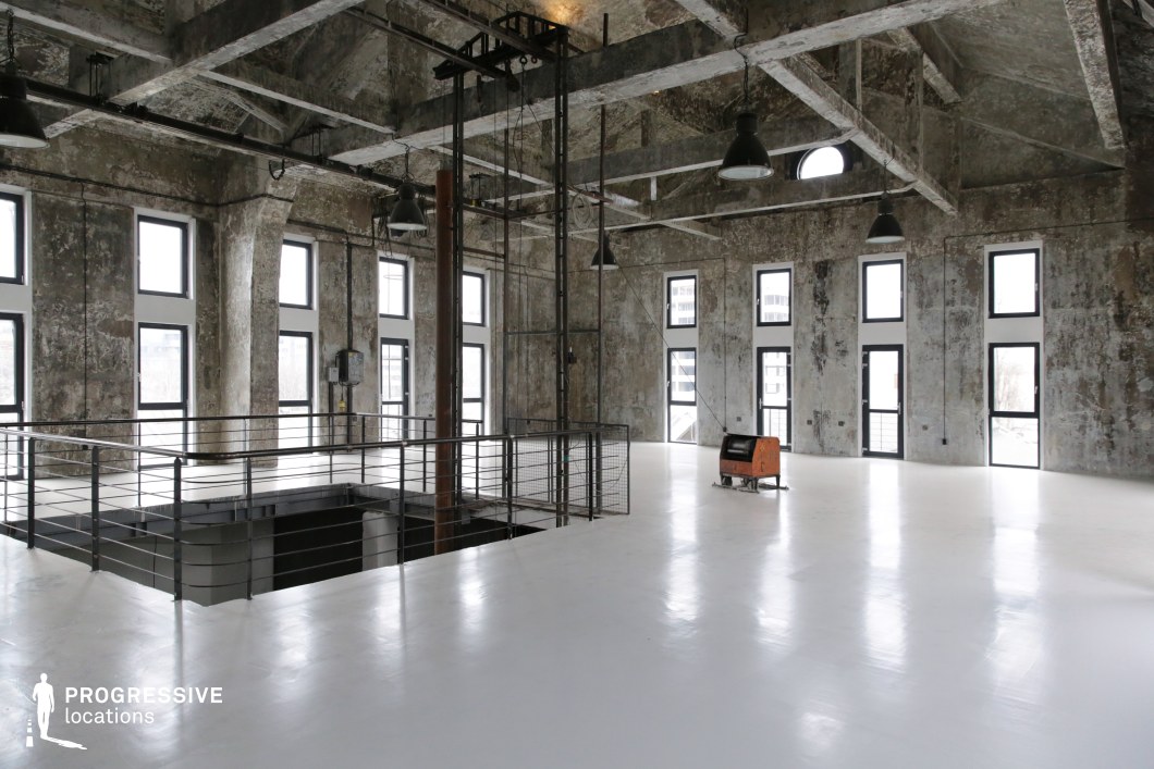  This spacious industrial loft features a striking contrast between its raw concrete walls and ceiling and its sleek, polished white flooring. Large, evenly spaced windows allow abundant natural light to enter, creating a bright yet rugged atmosphere. The high ceiling, exposed beams, and hanging industrial lamps enhance the open feel, while a central metal railing surrounds a descending staircase, adding depth to the composition. The space is minimal, with only a few scattered elements reinforcing its industrial past. This location is ideal for filming contemporary dramas, fashion shoots, or artistic productions, providing a versatile backdrop for both gritty and modern aesthetics.