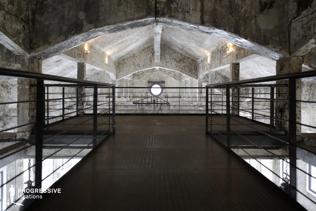 This industrial-style loft features a raised metal walkway flanked by sleek black railings, leading toward a circular window framed by a series of geometric, arched concrete beams. The raw, textured concrete walls and ceiling exhibit a weathered, unfinished look, contrasting with the modern metal elements. Soft lighting highlights the architectural depth, emphasizing the repeating patterns of the arches. The combination of old and new elements makes this an ideal filming location for futuristic, dystopian, or high-fashion productions, offering a visually rich setting that blends industrial grit with a minimalist, modern aesthetic.