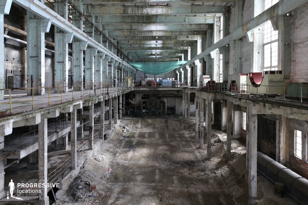 A massive abandoned industrial hall with towering concrete pillars and large multi-paned windows that allow natural light to filter through the dust-laden air. The structure features multiple levels of deteriorating walkways and railings, with remnants of machinery still in place. The floor is stripped bare, revealing dirt, rubble, and structural remnants, emphasizing its state of decay. The washed-out blue and white walls are weathered, showing cracks, stains, and rusted metal fixtures. A green safety netting hangs from the ceiling, adding a subtle industrial element. This vast, cavernous space is ideal for cinematic settings requiring a dystopian, post-apocalyptic, or historical industrial atmosphere.