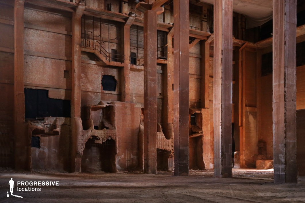 This vast, cavernous industrial space features towering rust-colored concrete columns supporting a multi-level brick facade, marked by decay and crumbling sections. The walls exhibit deep recesses, broken openings, and exposed steel reinforcements, adding to the raw, unfinished aesthetic. Shadows cast by the massive vertical supports create a dramatic contrast against the warm, earthy tones of the structure. The scale and industrial deterioration make this an ideal setting for dystopian, sci-fi, or historical thriller productions, serving as a forgotten manufacturing plant, an underground bunker, or a post-apocalyptic ruin.