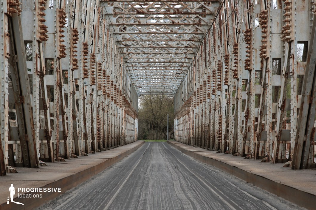 This long, rust-covered iron bridge features an intricate framework of metal beams and bolts, forming a tunnel-like passageway with a striking sense of depth. The corroded surfaces and peeling paint add to the weathered, abandoned aesthetic, while the worn asphalt road running through the center emphasizes its former use. The repetitive structural elements create a dramatic visual rhythm, making this location ideal for filming suspenseful, action-packed, or post-apocalyptic sequences. Perfect for chase scenes, confrontations, or desolate road trip settings, the bridge evokes themes of isolation, decay, and transition.