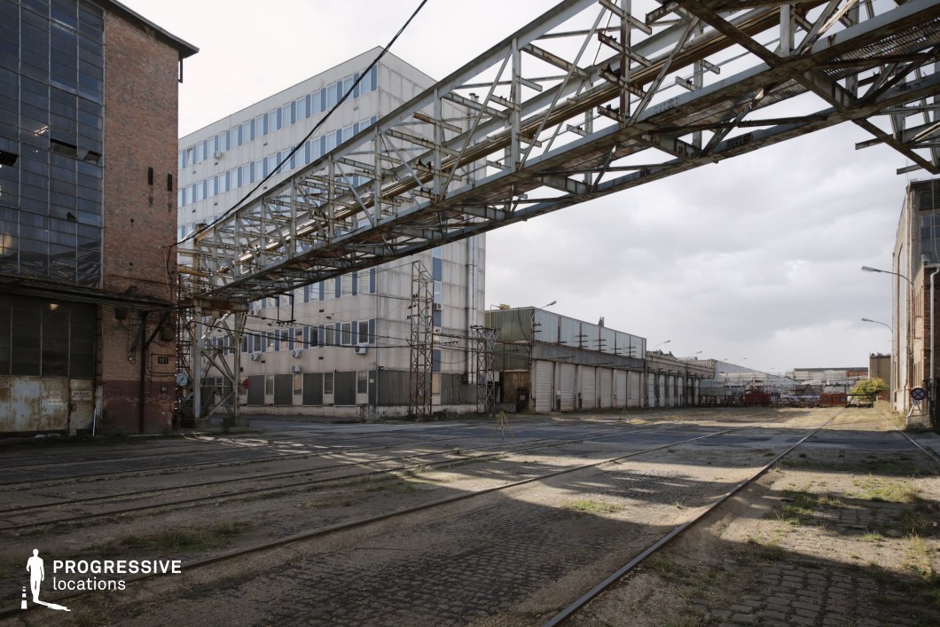 This expansive industrial yard features a rusted overhead metal walkway spanning between aging factory buildings, creating a striking visual element of decay and past functionality. The ground is lined with disused railway tracks embedded in cracked cobblestone, adding to the location’s gritty, abandoned atmosphere. In the background, large storage and manufacturing buildings with worn facades and faded signage contrast with a more modern office-like structure, reinforcing the blend of past and present industry. This location is ideal for filming dystopian, crime, or action sequences, providing an open yet structured setting suitable for chase scenes, covert operations, or industrial espionage narratives.