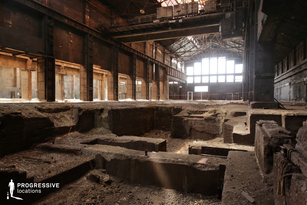 This vast, decayed industrial hall features towering steel columns, rusted metal beams, and a skeletal roof structure that allows diffused light to filter through shattered window panes. The floor is partially excavated, revealing deep, irregular concrete trenches that add a sense of desolation and structural decay. The walls are marked by years of grime and corrosion, with faded industrial remnants hinting at the building’s past functionality. A large overhead crane looms above, reinforcing the abandoned factory aesthetic. This location is ideal for filming dystopian, sci-fi, or action sequences, offering a gritty, post-apocalyptic setting with dramatic depth and scale.