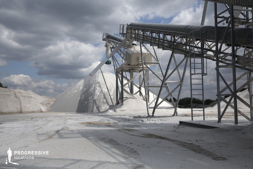 This large-scale mining or processing site features towering conveyor structures and metal frameworks, set against massive white mineral piles. The network of ladders, beams, and industrial chutes suggests an active material processing operation. The stark contrast between the bright ground and the metallic machinery creates a visually striking industrial landscape. The open space and geometric patterns of the structures provide a dynamic backdrop, making this location ideal for filming dystopian, sci-fi, or action sequences, as well as documentary-style industrial footage emphasizing large-scale production and extraction.