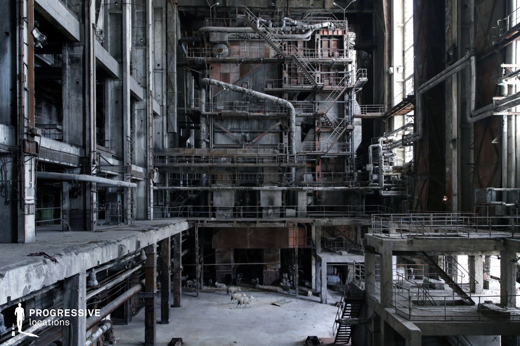 This massive industrial space features towering concrete pillars, rusted steel platforms, and an intricate network of aged pipes and staircases, creating a visually imposing mechanical labyrinth. The vast, multi-level structure is lined with corroded railings and catwalks, while large, grime-covered windows allow minimal light to penetrate the cavernous interior. The layers of decay, exposed metal framework, and weathered surfaces contribute to a dystopian, post-industrial atmosphere. This location is highly cinematic and well-suited for sci-fi, cyberpunk, or post-apocalyptic productions, serving as a decommissioned power plant, an abandoned factory, or a secret underground facility.