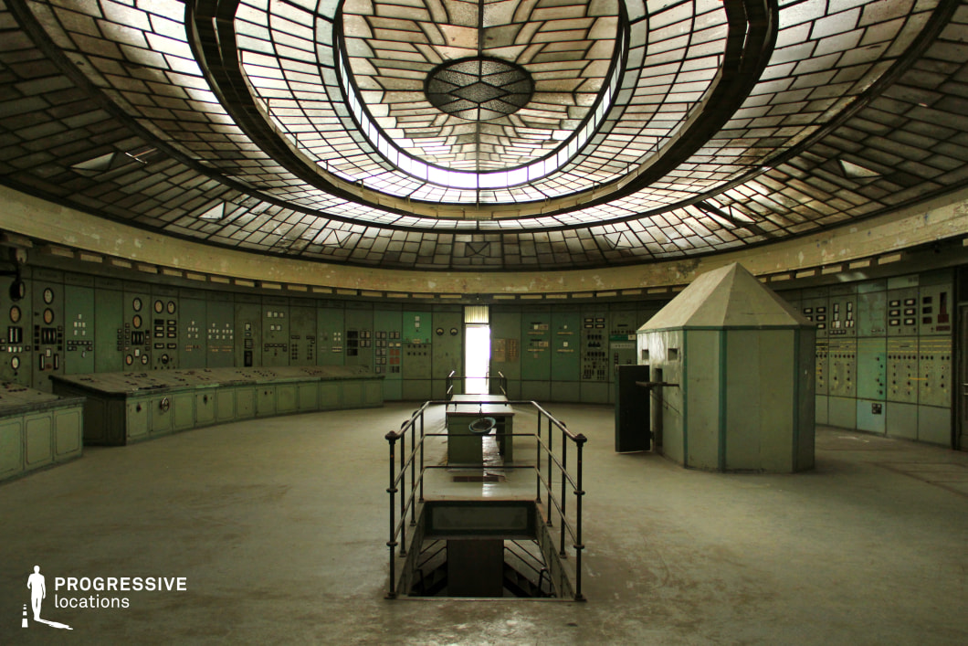 This large, circular control room features an impressive stained-glass ceiling with a geometric pattern that diffuses soft, atmospheric light into the space. The surrounding walls are lined with aged green control panels covered in analog dials, switches, and meters, reinforcing a vintage industrial aesthetic. At the center, a raised metal walkway leads to a recessed opening in the floor, adding a sense of mystery and function to the space, while a polygonal control booth stands nearby, hinting at past operational significance. The worn-out surfaces and peeling paint suggest abandonment, making this location ideal for dystopian, sci-fi, or historical thriller productions, serving as a decommissioned power facility, a secret government bunker, or a retro-futuristic command center.