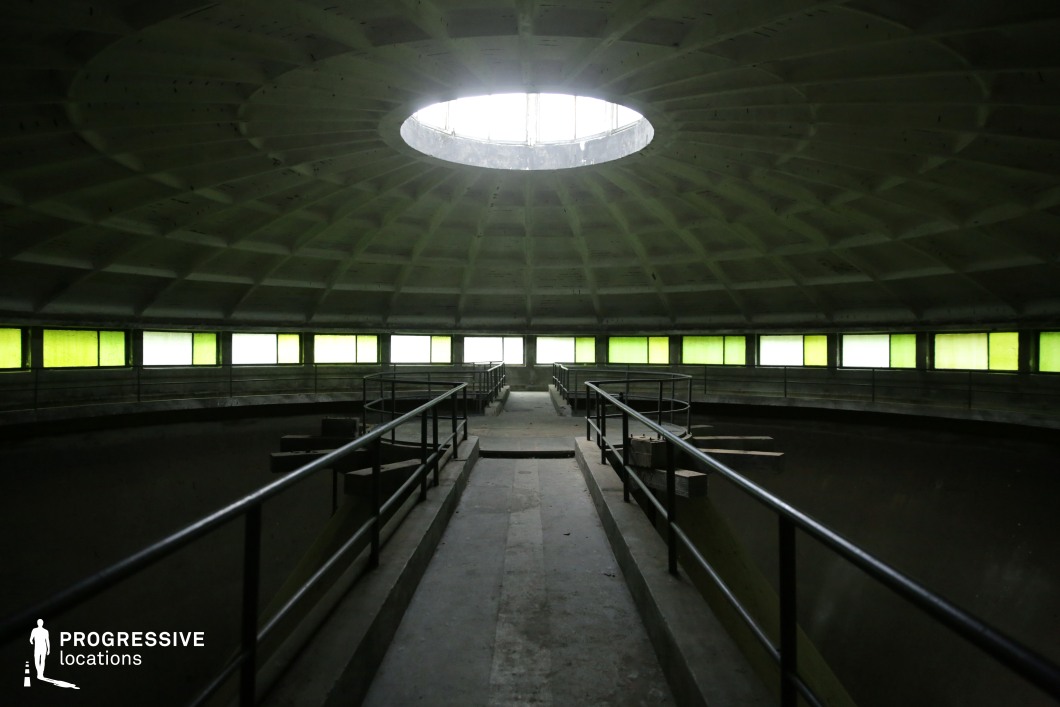 This striking circular structure features a domed concrete ceiling with a radial pattern, converging at a large central skylight that casts diffused light into the space. The perimeter is lined with green-tinted rectangular windows, creating an eerie glow that contrasts with the stark industrial design. A narrow concrete walkway with metal railings extends into the center, adding depth and symmetry to the composition. The empty, cavernous interior and geometric patterns make this location ideal for sci-fi, dystopian, or psychological thriller films, offering a futuristic or abandoned high-security facility aesthetic with strong visual impact.