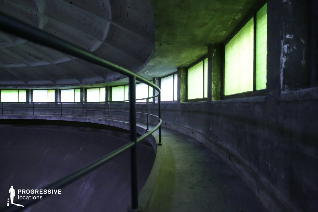 This circular concrete structure features a narrow, elevated walkway lined with metal railings, wrapping around a deep, recessed interior. A series of green-tinted windows casts an eerie glow, enhancing the space’s cold, industrial aesthetic. The curved ceiling and rough-textured walls emphasize the geometric precision of the architecture, while the repetitive pattern of the windows and railings create a hypnotic visual effect. This location is ideal for sci-fi, dystopian, or psychological thriller films, offering a futuristic yet decayed setting that evokes secrecy, surveillance, or confinement.