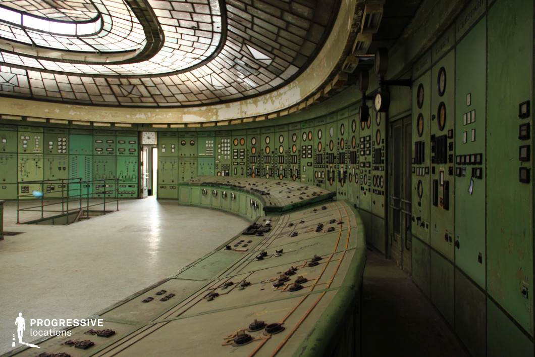 This abandoned control room features an expansive, curved console covered in dials, switches, and analog gauges, evoking a mid-20th-century industrial aesthetic. The walls are lined with green metal panels, densely packed with instrumentation, reinforcing a retro-futuristic, almost dystopian atmosphere. A striking glass ceiling with a geometric pattern diffuses natural light into the space, highlighting the peeling paint and worn surfaces that suggest long-term disuse. The room's spacious, symmetrical layout and central control desk make it visually compelling for cinematography, ideal for sci-fi, thriller, or historical films requiring an old industrial setting, a secretive command center, or a decommissioned power plant.
