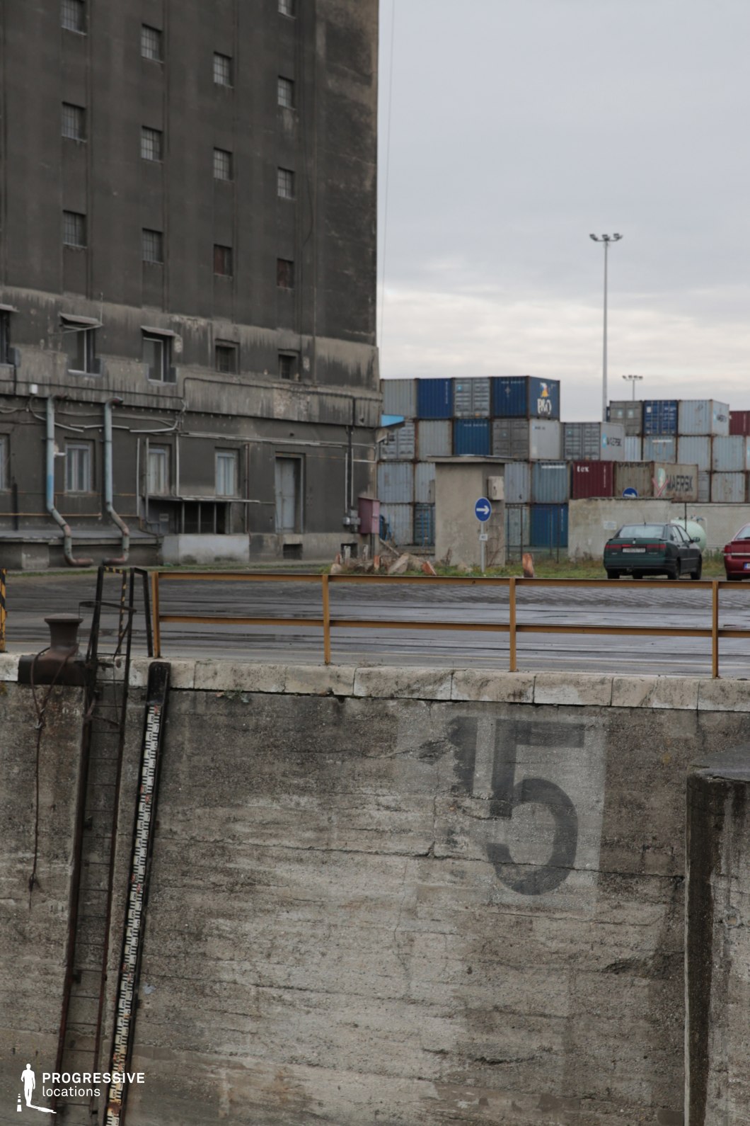 The image captures a gritty industrial dockside with a weathered concrete wall in the foreground, marked with a large, faded number “15.” A rusted metal ladder and a mooring bollard emphasize the location’s maritime function. Behind the barrier, a wide asphalt road with a simple metal railing separates the dock from an aged, dark grey industrial building, which features small, barred windows and exposed piping, contributing to a utilitarian and worn aesthetic. In the background, a towering stack of shipping containers in various muted colors, including blue, red, and grey, adds depth and scale, reinforcing the setting’s role as a working port. The raw textures of concrete, metal, and asphalt create an industrial, somewhat dystopian atmosphere. This location is ideal for filming gritty urban dramas, crime thrillers, or post-apocalyptic scenarios, offering a stark, utilitarian backdrop with a strong sense of isolation and decay.