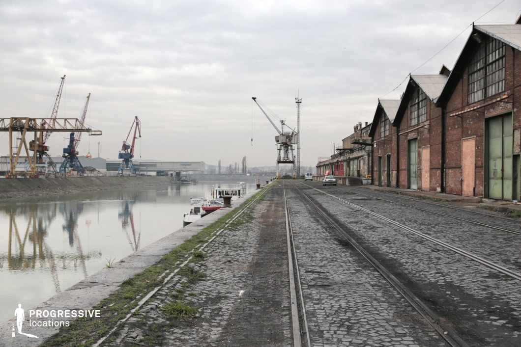 This expansive dockside setting features a long row of aging brick warehouses with large green metal doors lining a cobblestone street embedded with old railway tracks. The waterfront is lined with cranes and industrial equipment, reflecting in the calm water, while small boats are moored along the edge. The muted color palette and weathered surfaces contribute to a gritty, utilitarian aesthetic. This location is ideal for crime dramas, neo-noir, or thriller productions, serving as an industrial shipping hub, an abandoned port district, or a clandestine meeting point.