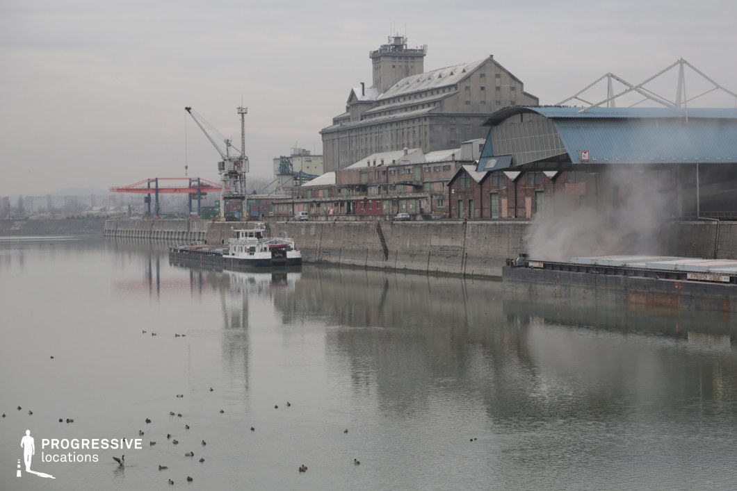 The image showcases an industrial river port featuring a mix of old and modern infrastructure. A large, multi-story concrete grain silo with a weathered facade dominates the background, accompanied by rows of aged brick warehouses with pitched roofs and metal reinforcements. A dockside crane and container-handling equipment indicate ongoing logistical operations, while moored cargo vessels and a barge emitting steam or smoke enhance the gritty industrial atmosphere. The river’s still waters reflect the structures, adding depth to the composition. This location is ideal for filming crime, thriller, or noir-style productions, as well as historical or industrial-themed scenes requiring an authentic, raw, and slightly dystopian setting.