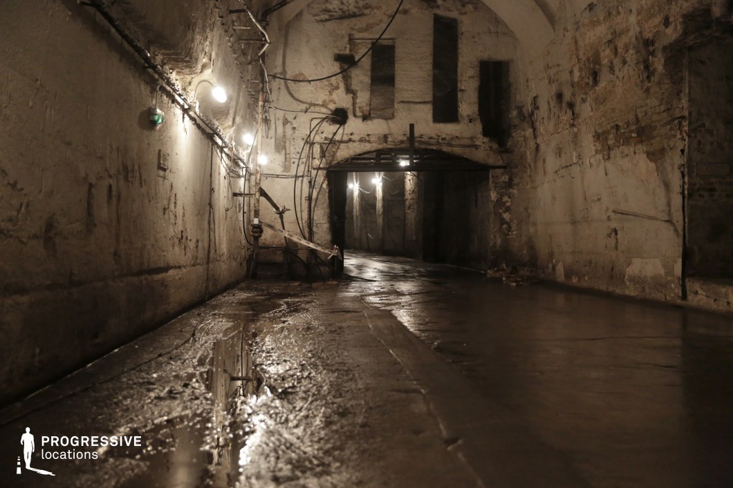 This dimly lit underground passage features rough, aged concrete walls and a curved ceiling, evoking a sense of decay and abandonment. The wet floor, with visible puddles and slick surfaces, reflects the overhead industrial-style lights that cast uneven illumination, emphasizing the tunnel’s depth and the rugged texture of the walls. Exposed pipes and electrical wiring run haphazardly along the structure, adding an industrial, almost forgotten atmosphere. A darkened opening in the distance suggests an extension of the passage, enhancing the sense of mystery. This location is ideal for filming tense chase sequences, horror or thriller scenes, or dystopian narratives, providing a confined, atmospheric setting with a strong visual impact.
