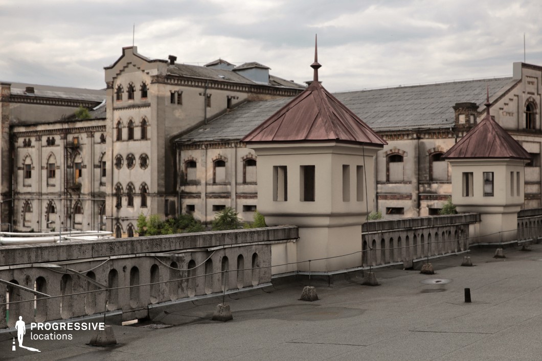 This expansive rooftop setting features a weathered concrete parapet adorned with arched cutouts and small guardhouse-style watchtowers with pointed copper roofs, evoking a fortified, institutional aesthetic. The aged main building in the background, with its gothic-inspired arched windows, circular openings, and faded stone facade, suggests a former industrial, correctional, or military function. The mix of architectural elements, from the fortress-like turrets to the grand but deteriorating structure, creates a sense of history and mystery. This location is ideal for filming tense escape sequences, espionage thrillers, or historical dramas, providing a dramatic, elevated vantage point with a sense of isolation and intrigue.