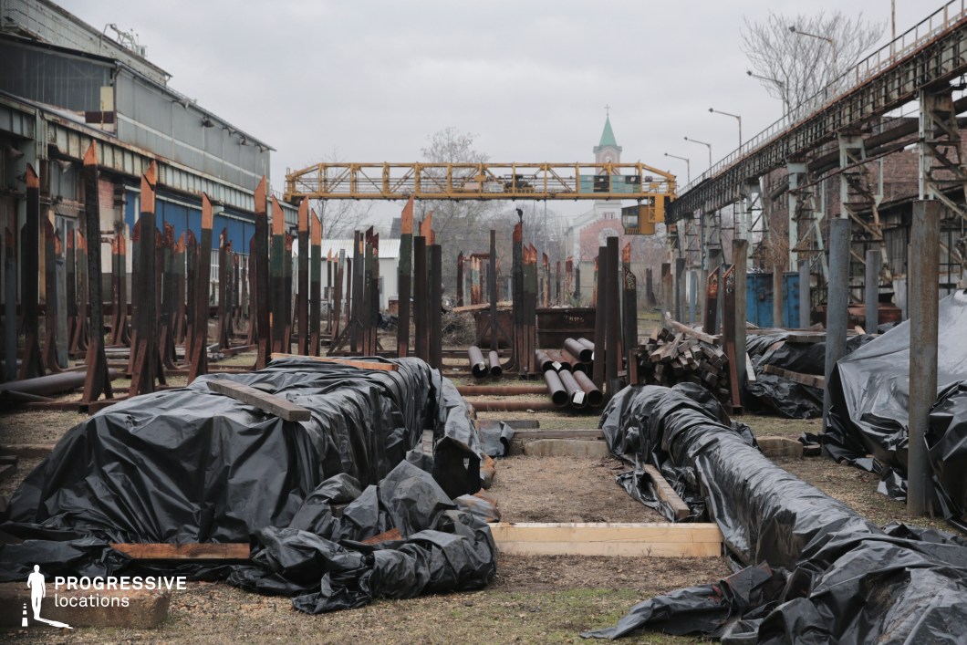 A desolate industrial yard filled with rusted metal beams, scattered pipes, and tarpaulin-covered materials, evoking a sense of abandonment and decay. The scene is framed by aged factory buildings with weathered metal sidings and overhead conveyor systems, adding to the industrial atmosphere. A yellow gantry crane stretches across the space, hinting at a past of heavy-duty manufacturing. In the background, a church steeple with a green roof emerges, juxtaposing the industrial remnants with historical architecture. This location offers a visually compelling setting for film productions needing a raw, gritty, and dystopian environment.