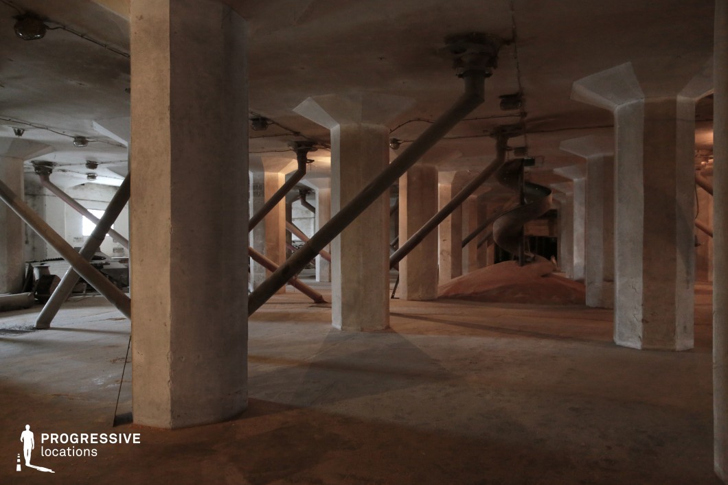 This expansive industrial space features thick concrete columns supporting a low ceiling, with metal reinforcement beams crisscrossing the area, giving it a functional and rigid geometric structure. Exposed pipes and grain chutes suggest the remnants of an old milling or storage facility, with piles of residual material settled on the floor. The dim lighting and earthy tones add to the atmosphere of abandonment and industrial decay. This location is ideal for filming historical, thriller, or dystopian scenes, providing a raw, mechanical backdrop suited for factory settings, underground bunkers, or clandestine operations.