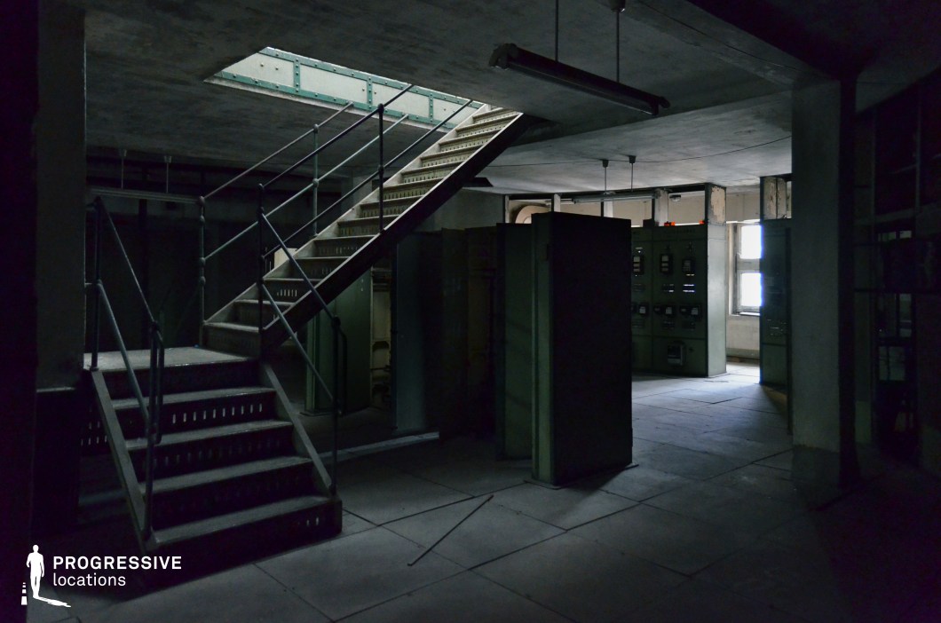 This dimly lit industrial control room features aged metal cabinets, control panels with analog dials, and an exposed concrete ceiling, evoking a sense of decay and secrecy. A metal staircase with railings leads to an upper level through an opening in the ceiling, partially illuminated by a skylight that casts a faint glow into the room. The walls show signs of wear, while scattered objects and darkened corners add to the unsettling atmosphere. This location is ideal for filming psychological thrillers, espionage sequences, or dystopian narratives, providing a setting that conveys secrecy, isolation, and tension.