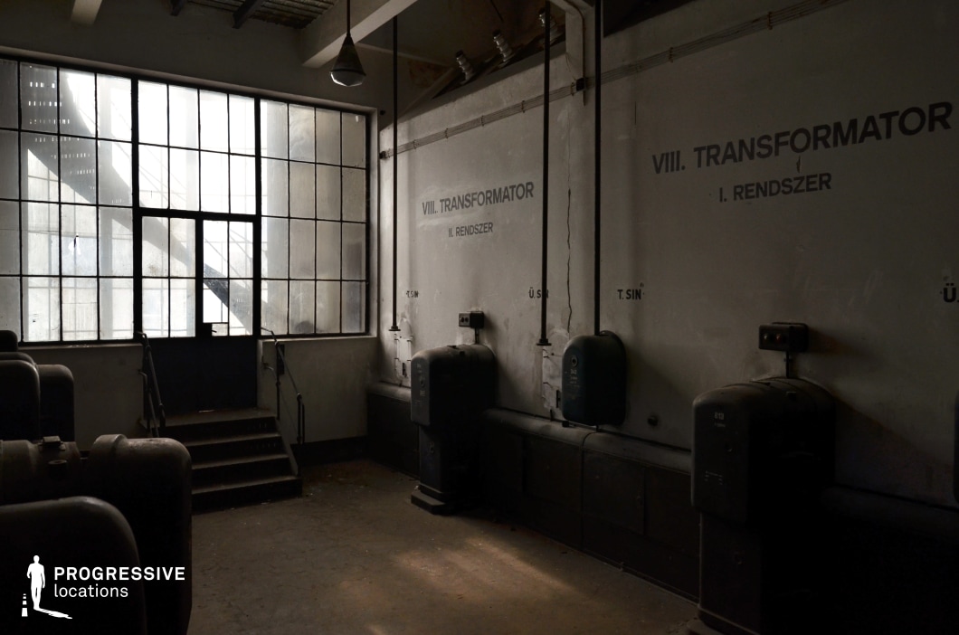 This dimly lit industrial space features aged concrete walls adorned with faded technical markings and mounted electrical units, contributing to its utilitarian and worn aesthetic. A large multi-paned window, partially obscured by grime, dominates one side, casting diffused light across the room and revealing an external metal staircase. A short flight of stairs with railings leads up to a heavy door, enhancing the sense of an enclosed, almost claustrophobic space. Exposed pipes and electrical conduits run along the walls, reinforcing the raw, functional feel of the setting. This location is ideal for filming tense, suspenseful scenes in sci-fi, crime, or industrial thriller genres, serving as a backdrop for clandestine meetings, power failures, or dystopian narratives.
