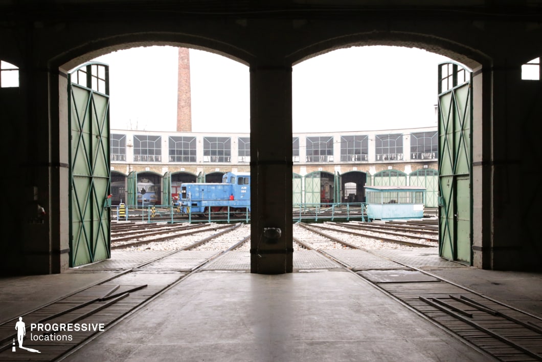 This industrial railway depot features a large arched entrance opening to multiple train tracks leading into a circular roundhouse. The metal-framed green doors, worn concrete flooring, and overhead structure create a strong sense of industrial heritage. A blue locomotive sits on the tracks, contrasting against the aged brick and glass-panelled depot, while a towering smokestack in the background adds depth to the composition. The symmetrical layout and open doors frame the view dynamically, making this location ideal for historical, adventure, or crime films requiring a vintage train setting, industrial-era aesthetics, or scenes involving railway operations.