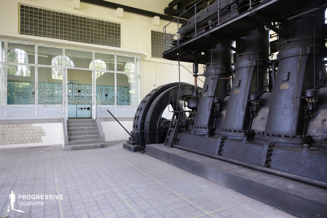 This historic industrial space features a massive black cast-iron turbine system dominating the foreground, with intricate pipes, bolts, and metalwork highlighting its early 20th-century engineering. Behind it, a glass-enclosed control room houses pale blue-green panels filled with analog dials and switches, offering a visually striking contrast between the mechanical power of the turbine and the structured order of the control space. The tiled floor and worn surfaces reinforce the sense of age and past industrial use. This location is ideal for historical, steampunk, or sci-fi productions, serving as an old power generation facility, a preserved industrial museum, or a secret experimental energy lab.