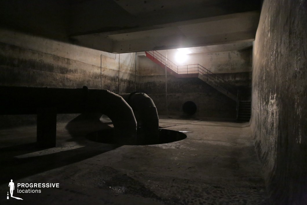This cavernous underground space features aged concrete walls stained with moisture and time, giving it a raw, abandoned appearance. Large, curved industrial pipes emerge from a dark opening in the floor, adding to the mechanical, utilitarian aesthetic. A metal staircase with railings leads to an upper level, illuminated by a single harsh light, casting deep shadows and enhancing the sense of depth. A circular tunnel opening in the far wall adds an element of mystery and intrigue. This location is ideal for filming tense, atmospheric scenes in sci-fi, horror, or dystopian settings, offering a confined, industrial backdrop with a sense of secrecy and unease.