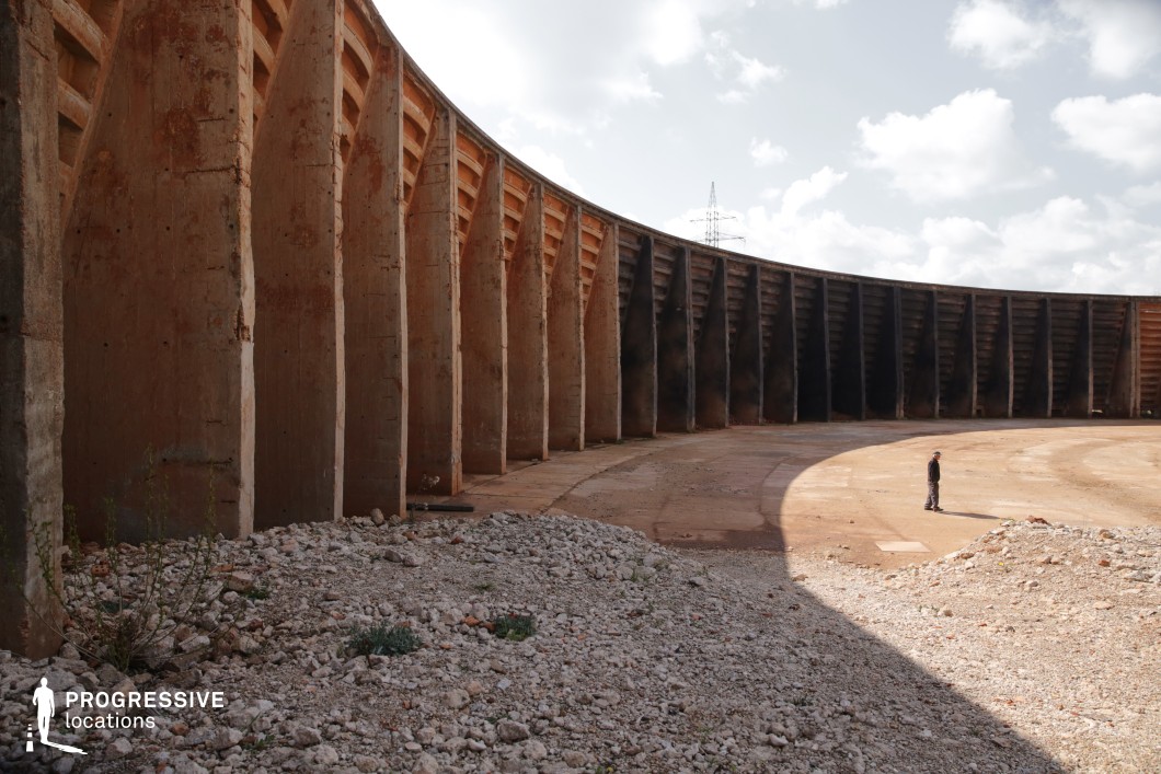 This vast circular industrial enclosure is lined with towering concrete walls reinforced by deep vertical supports, creating a monumental, almost monolithic structure. The ground is a mix of compacted earth and scattered rubble, adding to the desolate, utilitarian atmosphere. The stark contrast between light and shadow along the curved wall enhances the dramatic visual impact. This location is ideal for dystopian, sci-fi, or post-apocalyptic productions, serving as an abandoned storage facility, a decommissioned industrial zone, or a futuristic gladiatorial arena.
