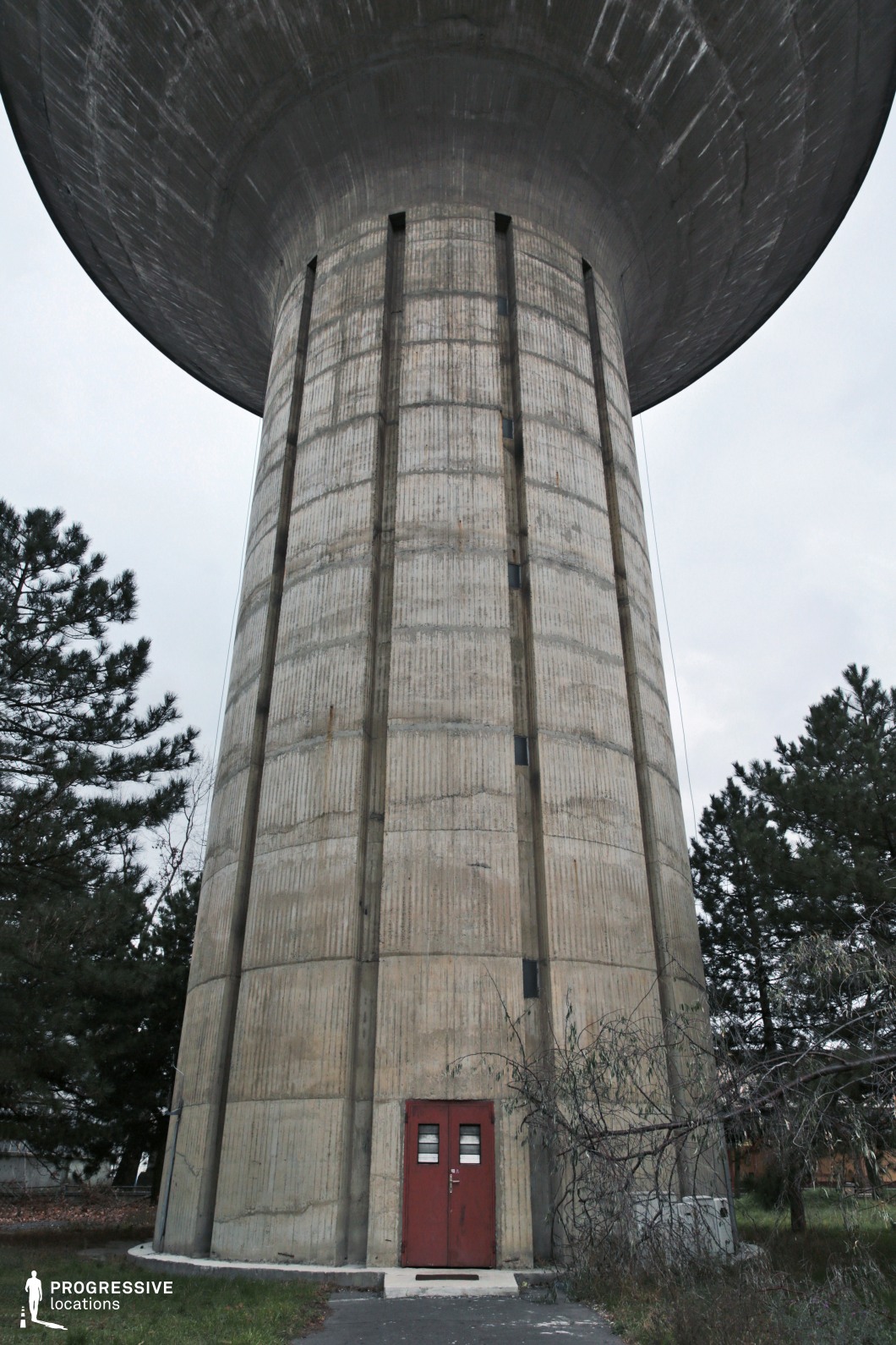 This towering concrete structure exemplifies brutalist architecture, with vertical ribbed patterns and a massive overhanging top. The imposing cylindrical form is accentuated by a small red metal door at its base, creating a stark contrast against the raw, weathered concrete. Narrow slit-like windows run vertically along the structure, adding to its monolithic presence. The surrounding area features patches of overgrown vegetation, reinforcing a slightly dystopian or abandoned aesthetic. This location is ideal for filming science fiction, dystopian, or thriller scenes, offering a visually striking and imposing setting that evokes themes of isolation, secrecy, or industrial power.