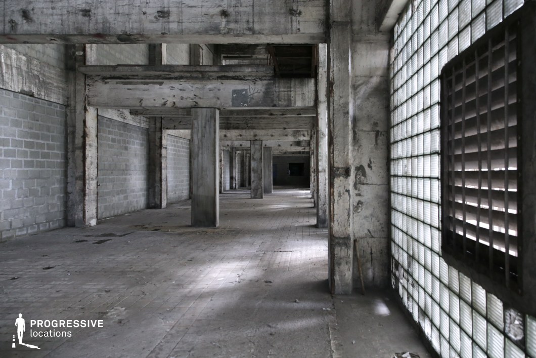 This long, desolate corridor features raw concrete beams, worn tiled flooring, and a repetitive column structure that creates a strong sense of depth and perspective. The right side is lined with textured glass block windows, partially covered by metal shutters, allowing diffused light to cast a cold, sterile glow across the space. The walls show significant wear, with peeling paint and exposed structural elements adding to the atmosphere of decay. The symmetrical alignment of pillars and the narrowing vanishing point make this location ideal for filming psychological thrillers, dystopian or post-apocalyptic scenes, or crime dramas requiring a confined yet visually striking industrial setting.
