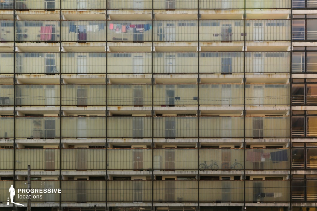 The image presents a repetitive, grid-like facade of an aging mid-century apartment block, where enclosed balconies with translucent glass panels create a uniform yet worn aesthetic. The muted yellowish tint of the glass, rust-streaked metal framing, and subtle signs of habitation give the structure a sense of functional brutalism. The rigid symmetry and lack of ornamentation emphasize the building’s utilitarian purpose, while small details, such as faint graffiti and laundry hanging behind the panels, add a human element. This location is ideal for dystopian films, social realism, or psychological thrillers, offering a stark, almost claustrophobic backdrop that conveys themes of conformity, isolation, or urban decay.