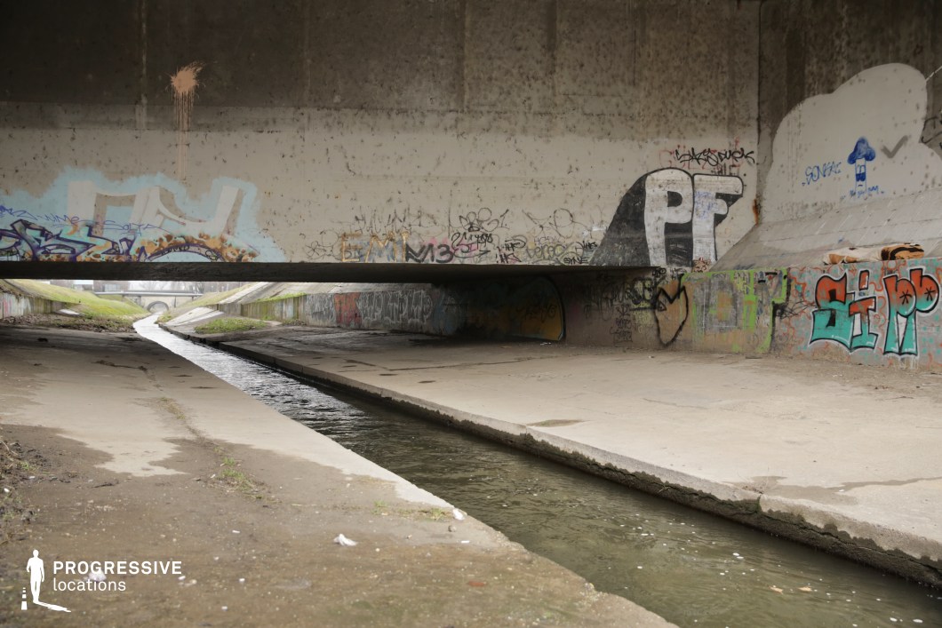 A raw and unfiltered urban landscape beneath a concrete overpass, where a narrow canal flows through a gritty, graffiti-covered underpass. The weathered walls are adorned with layers of spray paint, tags, and murals, adding to the space's raw, rebellious aesthetic. The low bridge creates a compressed, almost claustrophobic atmosphere, while the canal's reflective surface provides a subtle contrast to the rough, industrial surroundings. The distant opening toward the horizon hints at continuity beyond the underpass, making this setting ideal for film scenes requiring an urban, underground, or dystopian aesthetic.