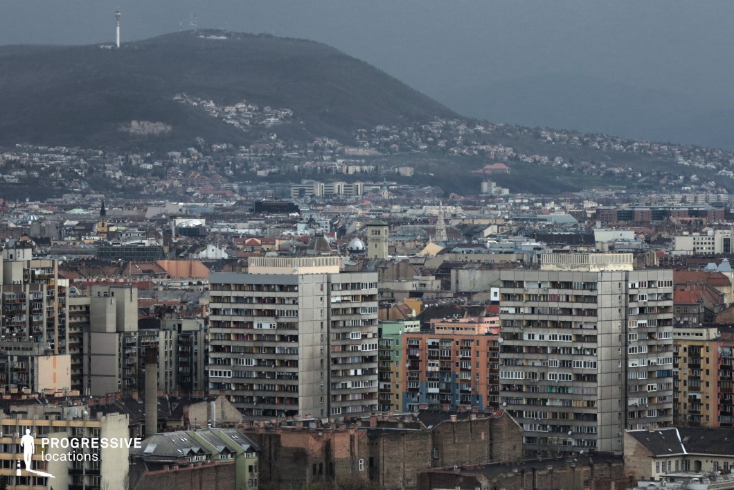 The image showcases a dense urban landscape dominated by large, brutalist-style residential buildings with repetitive concrete facades, long balconies, and rectangular windows. These towering structures contrast with the surrounding lower, older buildings that feature a mix of architectural styles, including ornate rooftops and colorful facades. In the background, rolling hills dotted with houses rise above the cityscape, adding depth and scale to the composition. The contrast between the rigid geometry of the high-rises and the more organic sprawl of traditional buildings creates a visually layered urban environment. This location is ideal for filming gritty urban dramas, dystopian sci-fi settings, or political thrillers, providing a visually compelling mix of modernist uniformity and historical texture.